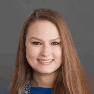 Smiling person with long brown hair wearing a blue top and multicolored scarf, posed against a plain gray background in a professional headshot.