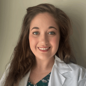 Smiling person with long brown hair wearing a white lab coat over a green floral-patterned top, posed against a plain light background. The attire and setting suggest a professional in audiology.