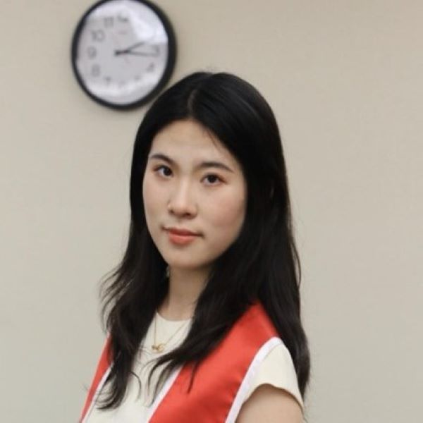Person with long black hair wearing a white outfit and a red and white sash, standing indoors in front of a plain wall with a clock. The individual is smiling, capturing a moment that suggests celebration or recognition.