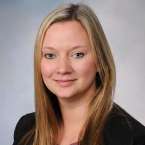 Professional portrait of a person with long, straight blonde hair, wearing a black blazer over a mauve top. They are facing the camera with a neutral expression against a soft blue and light gray gradient background, commonly used in formal or corporate photography.