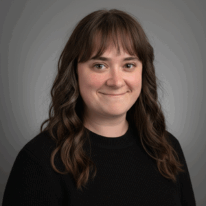 Portrait of a person with long, wavy brown hair wearing a black ribbed sweater, smiling slightly while facing the camera. The background is a plain gray gradient, giving the image a professional studio feel.