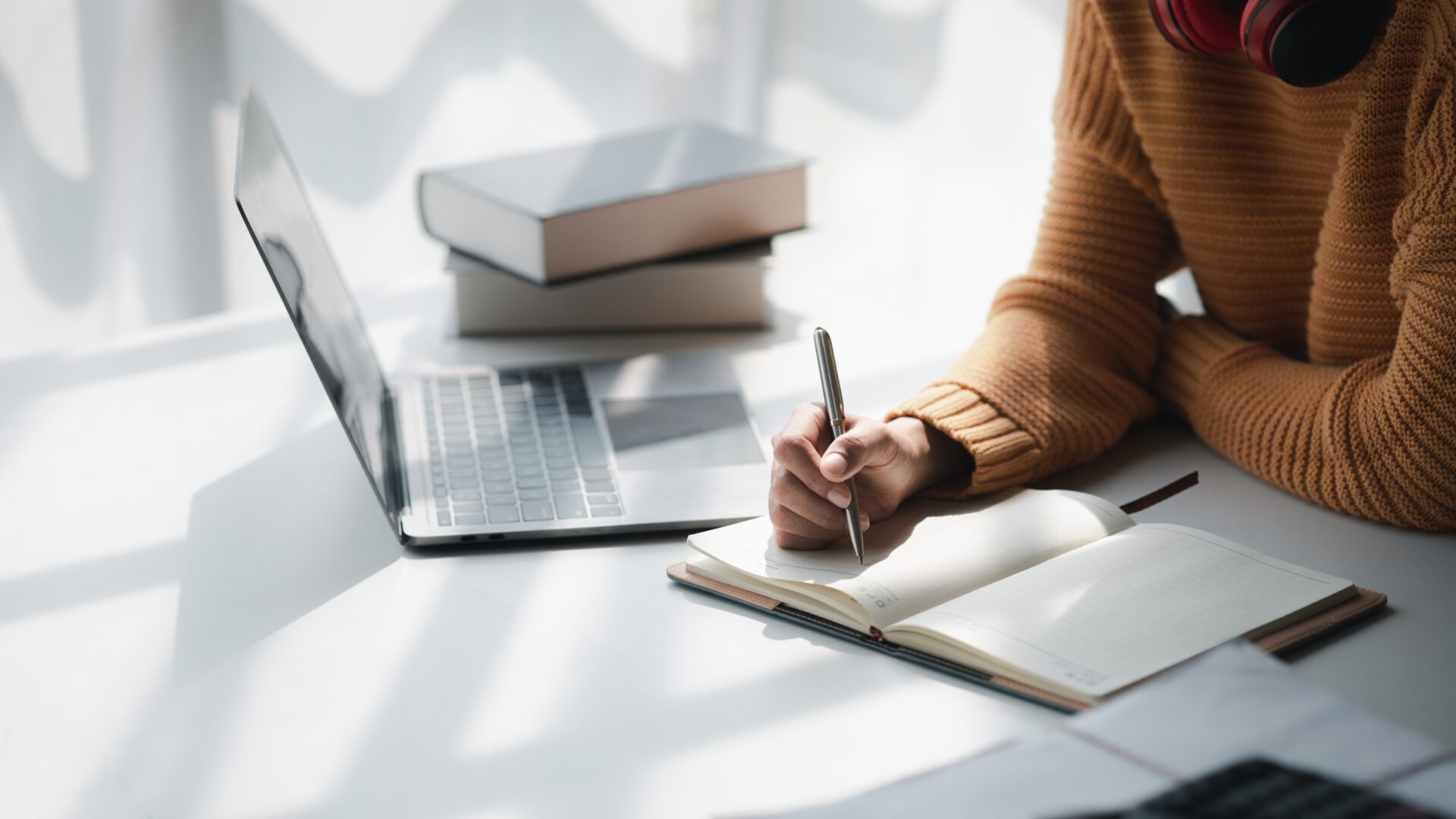 A person wearing a brown knitted sweater and red headphones sits at a desk, writing in an open notebook. The desk holds a laptop, a stack of books, and scattered papers, all illuminated by soft natural light from a nearby window with sheer curtains—creating a calm, focused study environment.