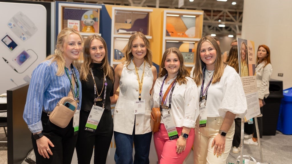 A group of five people stand together at a professional conference, smiling at the camera. They are wearing name badges and lanyards. Behind them are posters and display boards, including one that says “Meet Design Team!” with a photo of a person named Kristin. Other attendees and booths are visible in the background, creating a busy conference atmosphere.