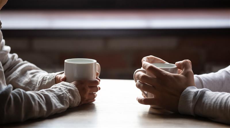 Two people sit across from each other at a wooden table, each holding a white mug with both hands. The scene is softly lit, with a blurred brick wall and window in the background, creating a warm, conversational atmosphere.