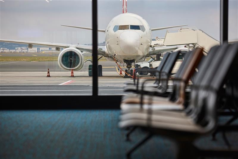 A commercial airplane is parked at an airport gate, viewed from inside the terminal through large windows. A jet bridge is connected to the aircraft, and empty rows of terminal seating are visible in the foreground.