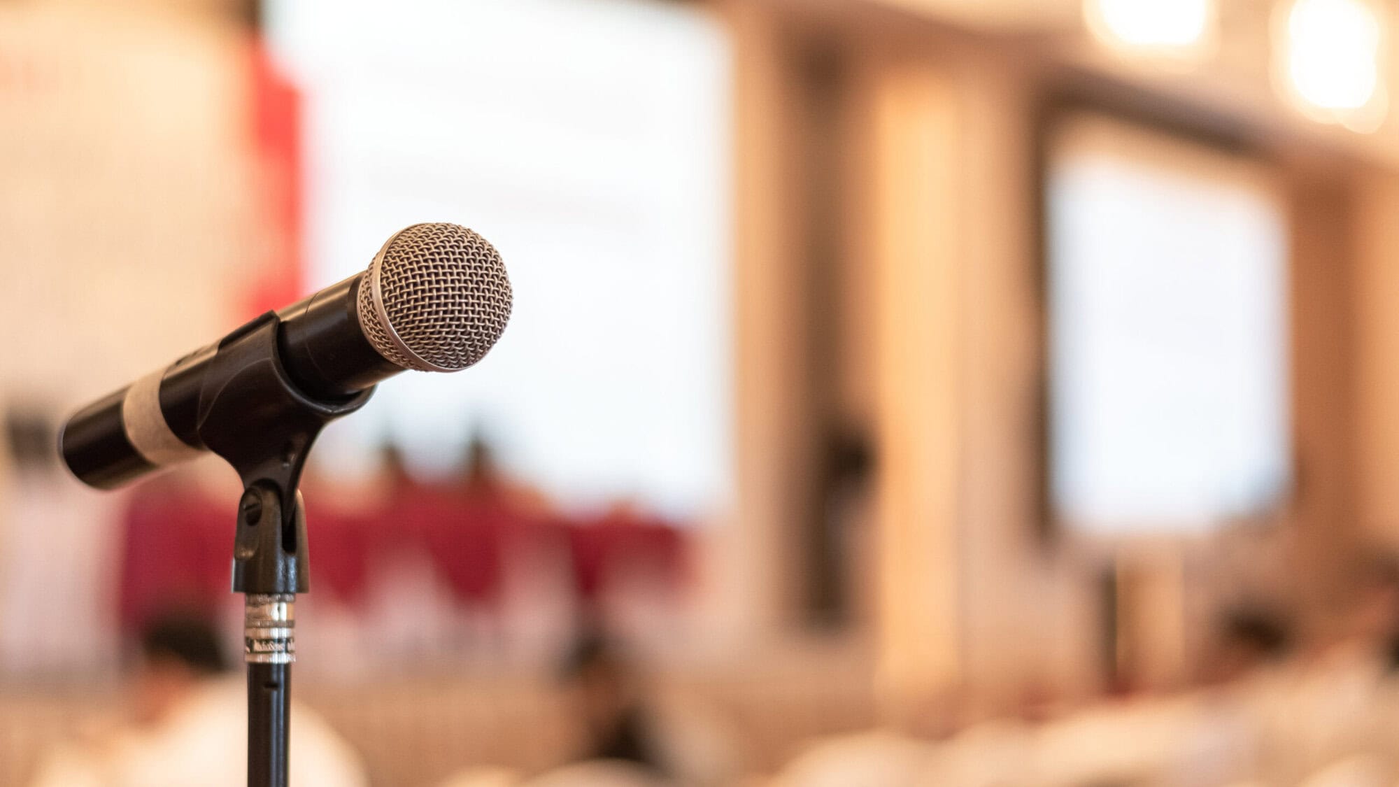 A close-up of a microphone on a stand, with a metallic mesh head and black body, positioned in front of a softly blurred indoor background that resembles a conference room or auditorium with warm lighting and indistinct figures.