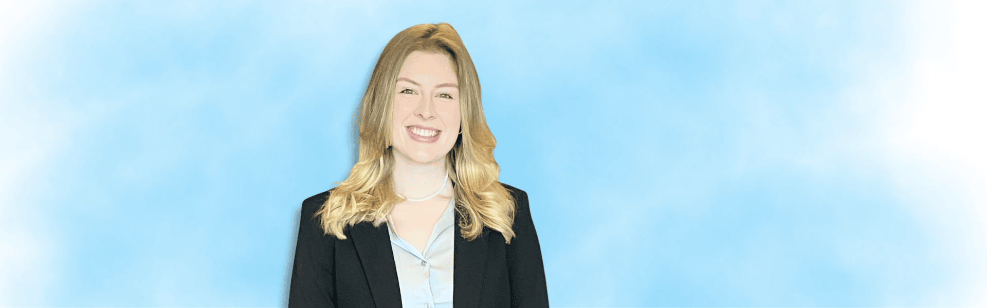 A professional portrait of a female individual with light-colored hair wearing a black blazer over a light blue shirt, standing against a soft light-blue background and facing the camera with a warm expression.