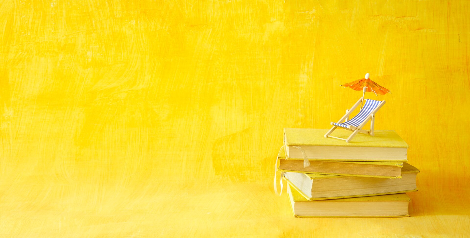 A small beach chair with a matching umbrella resting on top of a neatly stacked pile of books, all set against a bright yellow background.