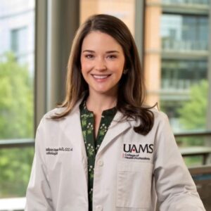 A woman wearing a white lab coat stands indoors near tall windows with greenery and a modern building visible outside. Her lab coat reads “Madison Haws Au.D., CCC-A Audiologist” on one side and “UAMS College of Health Professions” on the other. She wears a green patterned shirt underneath and smiles slightly in a bright, professional healthcare setting.