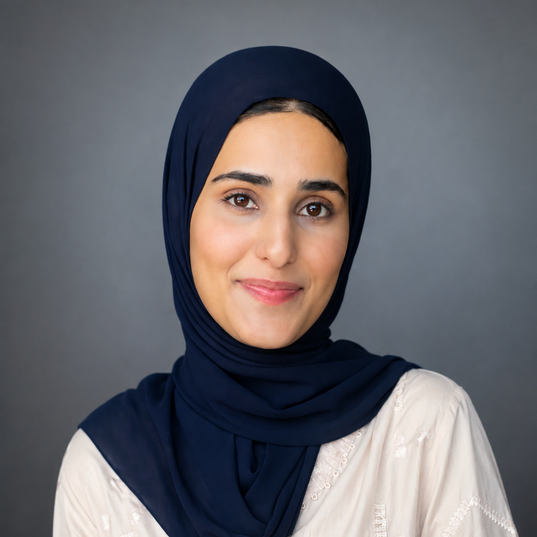 A woman wearing a dark navy hijab and a light-colored embroidered top poses against a plain gray background, looking directly at the camera with a calm, neutral expression.