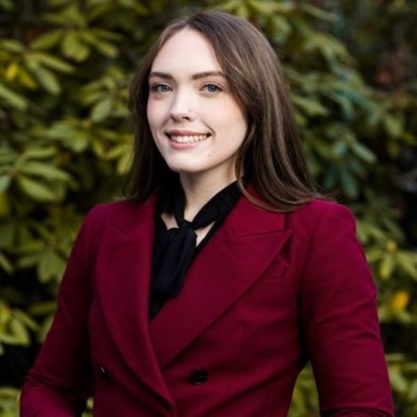 Person with long brown hair smiling at the camera, wearing a deep red blazer and a black scarf tied around the neck. They are standing outdoors in front of lush green foliage, suggesting a professional or formal portrait in a natural setting.