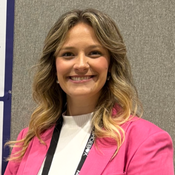 Here’s a polished alt text for the image: Alt Text: Person with shoulder-length wavy blonde hair standing in front of a gray fabric-covered background, smiling. They are wearing a bright pink blazer over a white top and a black lanyard with the text suggesting a professional or academic setting.