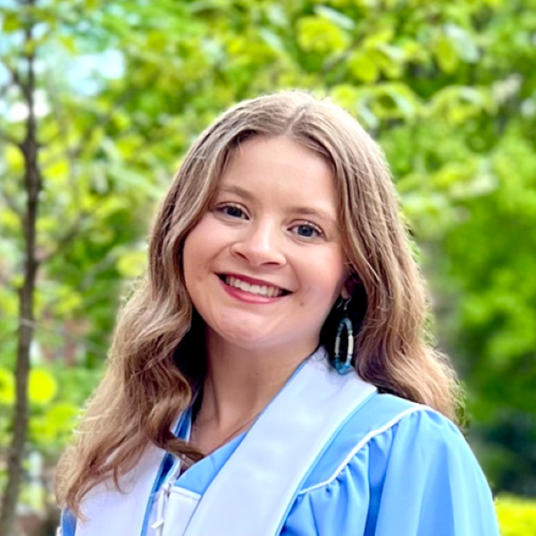 oung person with long, wavy brown hair wearing a light blue graduation gown and white stole, standing outdoors in front of green foliage. They are smiling at the camera, capturing a joyful moment of academic achievement.