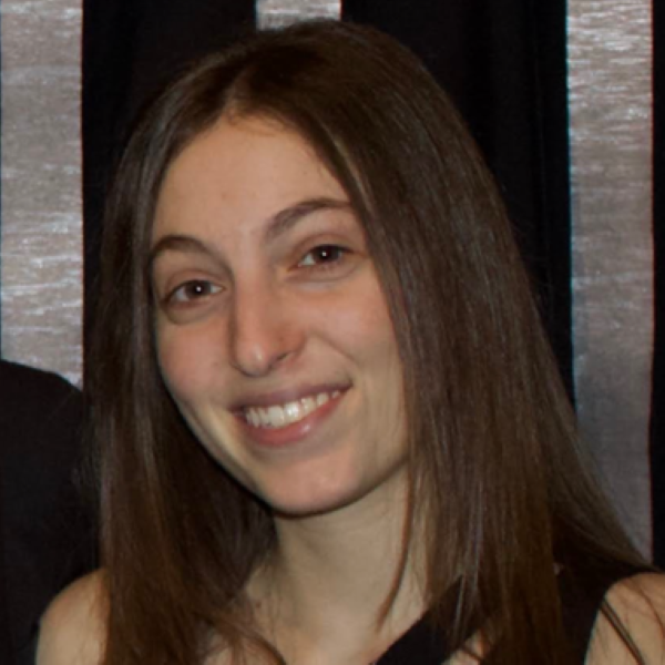 Person with long straight brown hair smiling at the camera, wearing a black sleeveless top. They are standing in front of a backdrop with alternating vertical black and silver stripes, suggesting a formal or semi-formal event setting.
